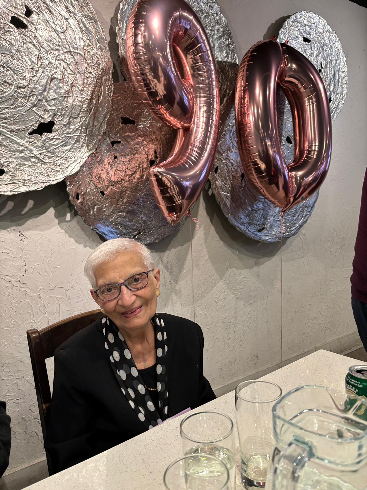 Maa celebrating her 90th birthday seated at a table with big balloons behind her